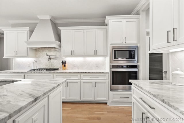 12113 S Fulton Ave E, Bixby, OK 74008 - kitchen with appliances with stainless steel finishes, custom exhaust hood, white cabinetry, light wood-style floors, and crown molding