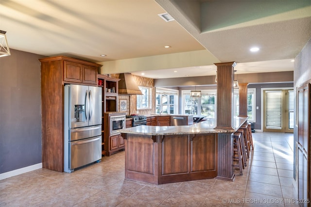 5378 W 450th Rd, Pryor, OK 74361 - kitchen featuring appliances with stainless steel finishes, ornate columns, light stone counters, wall chimney range hood, and a breakfast bar area