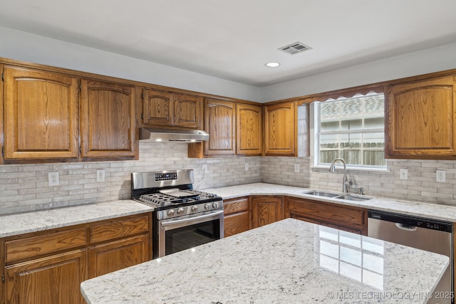 kitchen with appliances with stainless steel finishes, under cabinet range hood, a sink, brown cabinetry, and light stone countertops