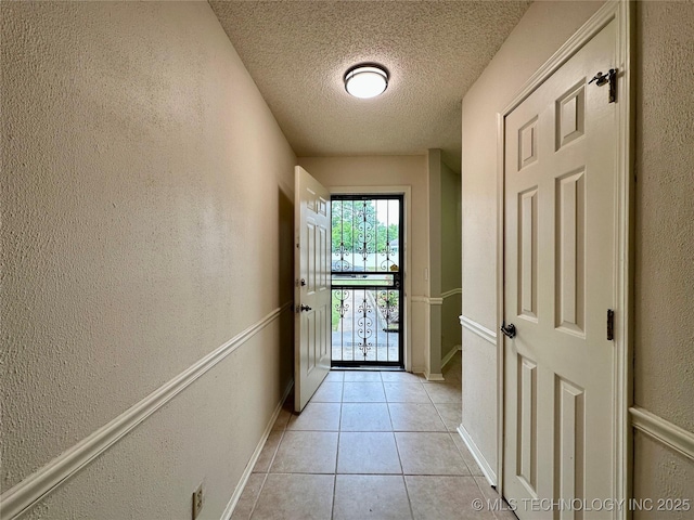 doorway with a textured ceiling, light tile patterned floors, a textured wall, and baseboards