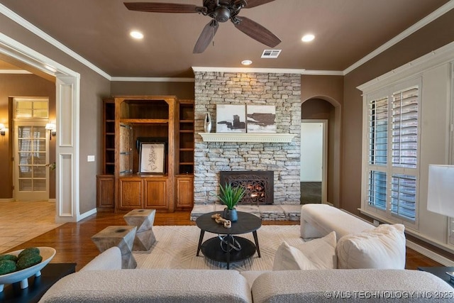 living room featuring arched walkways, crown molding, a ceiling fan, a stone fireplace, and wood finished floors