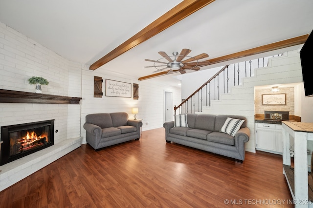 living area featuring a brick fireplace, stairs, dark wood-style floors, ceiling fan, and beam ceiling