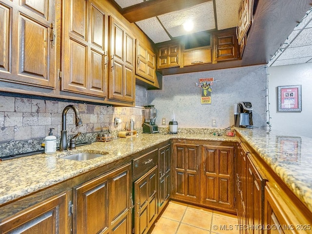 kitchen with a sink, tasteful backsplash, light stone countertops, light tile patterned floors, and brown cabinetry
