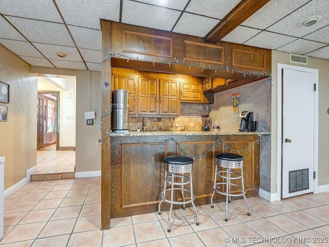 kitchen featuring light tile patterned flooring, a paneled ceiling, a peninsula, brown cabinetry, and backsplash