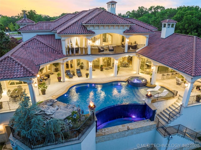 5879 N Eagle Summit Rd, Owasso, OK 74055 - back of house at dusk with a balcony, a pool with connected hot tub, a tile roof, a chimney, and an outdoor hangout area