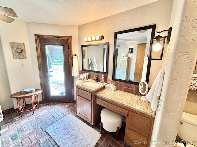 2102 N Peaceful Ln, Stigler, OK 74462 - bathroom with vanity, a textured wall, brick flooring, toilet, and ceiling fan