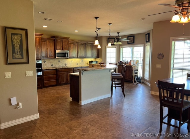 7400 E Prairie View Rd E, Kenefic, OK 74748 - kitchen with stainless steel microwave, a ceiling fan, a kitchen breakfast bar, backsplash, and an island with sink