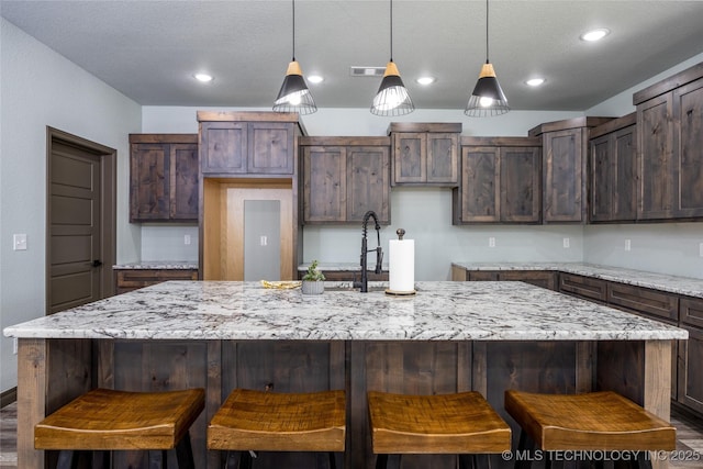 101 Dunn, Caddo, OK 74729 - kitchen featuring a textured ceiling, a center island with sink, light stone counters, decorative light fixtures, and dark brown cabinetry