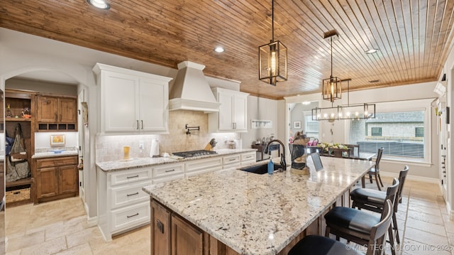 7920 N 147th E Ave, Owasso, OK 74055 - kitchen with custom range hood, wood ceiling, stainless steel gas stovetop, a center island with sink, and stone tile flooring
