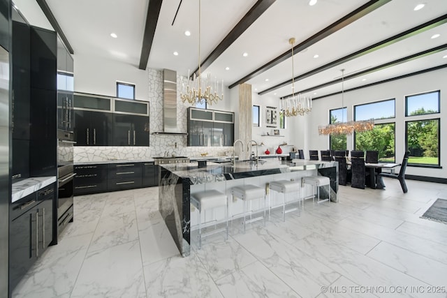 1226 W 109th Pl S, Jenks, OK 74037 - kitchen featuring wall chimney exhaust hood, dark cabinetry, a chandelier, beamed ceiling, and a kitchen breakfast bar