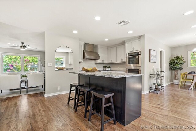 5861 S Hudson Pl, Tulsa, OK 74135 - kitchen with wall chimney range hood, double oven, a breakfast bar area, a center island, and a ceiling fan