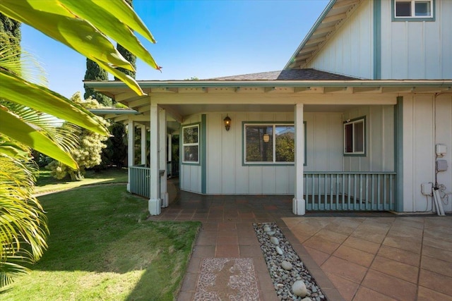 view of exterior entry with a patio, a lawn, board and batten siding, and roof with shingles