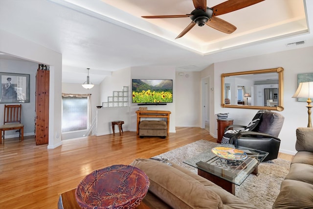 living area featuring light wood-type flooring, visible vents, baseboards, a raised ceiling, and ceiling fan