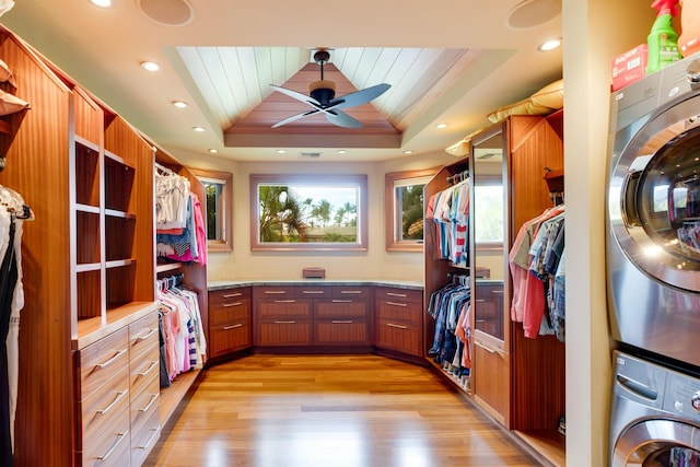 walk in closet with ceiling fan, stacked washer and clothes dryer, a tray ceiling, and light wood-type flooring