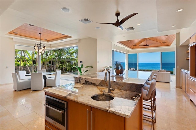 kitchen featuring light stone countertops, a raised ceiling, a kitchen island with sink, decorative light fixtures, and a water view