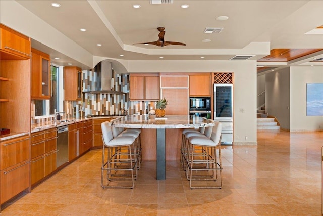 kitchen with light stone countertops, appliances with stainless steel finishes, wall chimney exhaust hood, a raised ceiling, and a center island