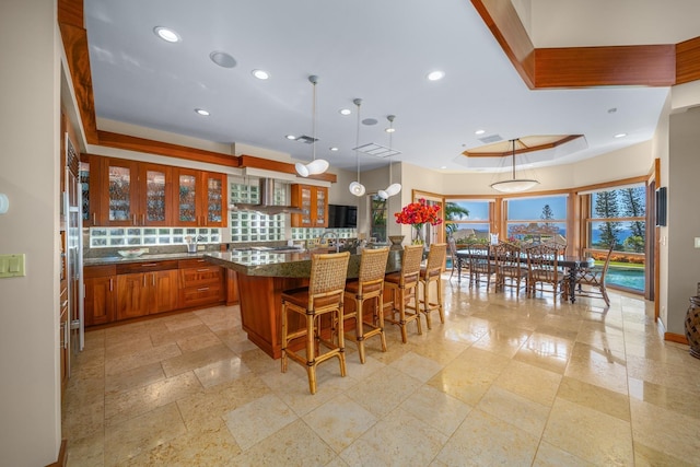 kitchen featuring a kitchen breakfast bar, a raised ceiling, wall chimney range hood, decorative light fixtures, and a kitchen island