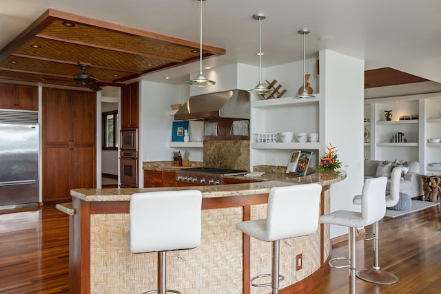 kitchen featuring wall chimney range hood, built in appliances, dark wood-type flooring, and hanging light fixtures