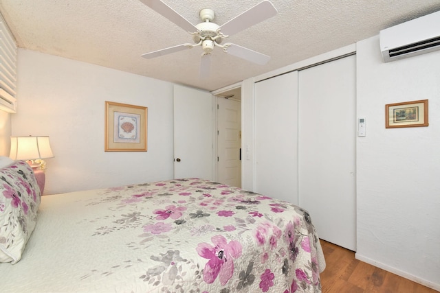 bedroom with a wall mounted air conditioner, a closet, wood-type flooring, ceiling fan, and a textured ceiling