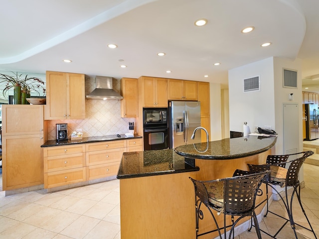 kitchen with wall chimney exhaust hood, dark stone countertops, a center island with sink, light tile patterned floors, and black appliances