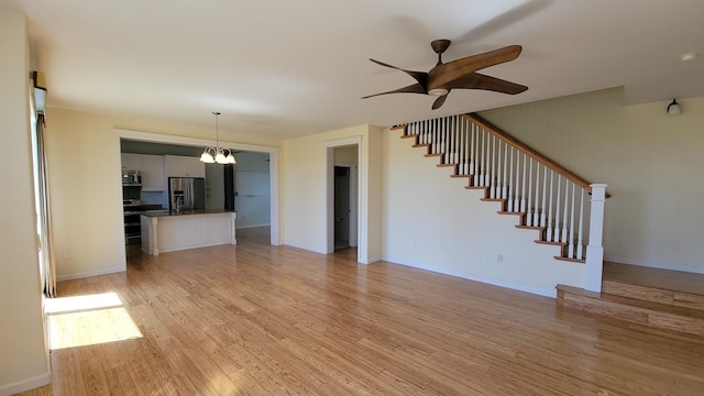 unfurnished living room with light wood-style floors, stairway, a chandelier, ceiling fan, and baseboards