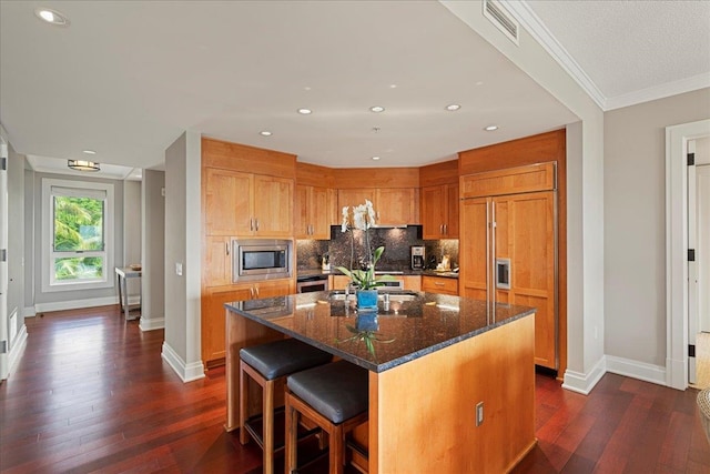 kitchen with dark wood-style flooring, visible vents, backsplash, brown cabinetry, and built in appliances