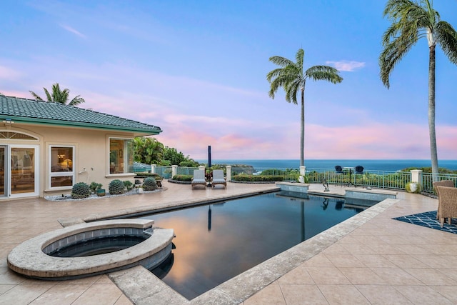 pool at dusk with an in ground hot tub, a water view, and a patio area