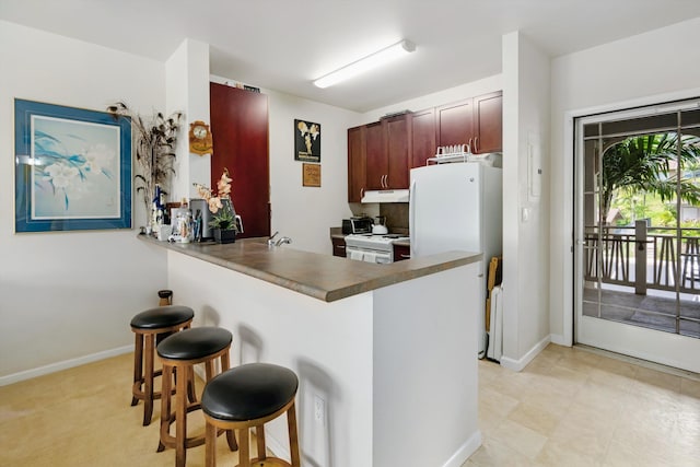 kitchen featuring a breakfast bar, white appliances, a peninsula, baseboards, and under cabinet range hood