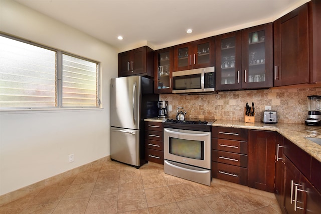 kitchen featuring stainless steel appliances, glass insert cabinets, backsplash, and light stone countertops