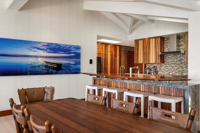 kitchen with decorative backsplash, kitchen peninsula, light hardwood / wood-style flooring, vaulted ceiling with beams, and wall chimney exhaust hood