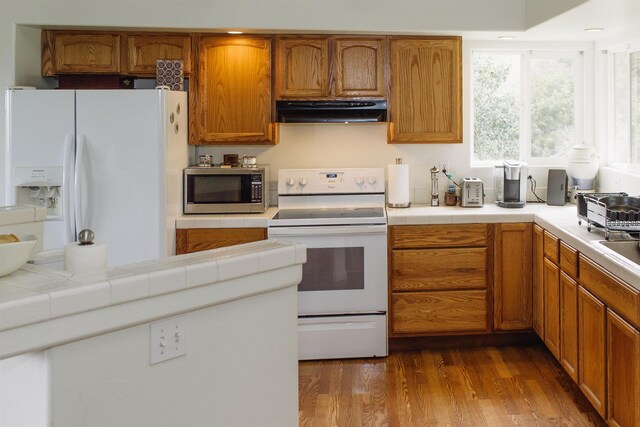 kitchen featuring under cabinet range hood, tile countertops, white appliances, dark wood-type flooring, and brown cabinetry