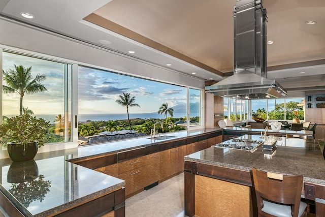 kitchen featuring brown cabinets, dark stone countertops, island exhaust hood, stainless steel gas stovetop, and a sink