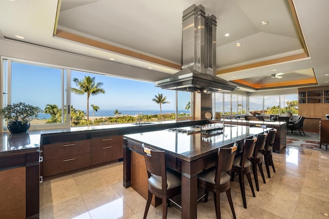 kitchen featuring a tray ceiling, a sink, dark stone countertops, gas cooktop, and island range hood