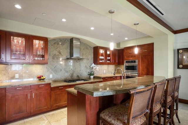 kitchen with wall chimney range hood, decorative light fixtures, decorative backsplash, a center island with sink, and appliances with stainless steel finishes