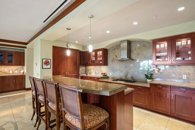 kitchen featuring wall chimney exhaust hood, decorative backsplash, an island with sink, decorative light fixtures, and stainless steel gas cooktop