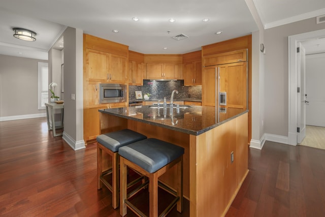 kitchen with dark wood-type flooring, dark stone counters, a kitchen breakfast bar, built in appliances, and an island with sink