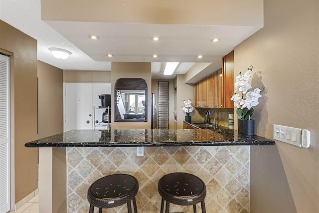 kitchen with backsplash, dark stone countertops, a peninsula, brown cabinetry, and a sink