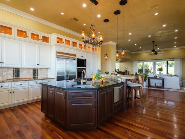 kitchen with built in appliances, a center island with sink, white cabinets, and ceiling fan with notable chandelier