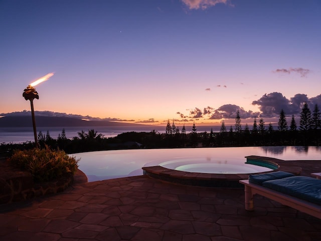 pool at dusk featuring an in ground hot tub, a water view, and a patio area