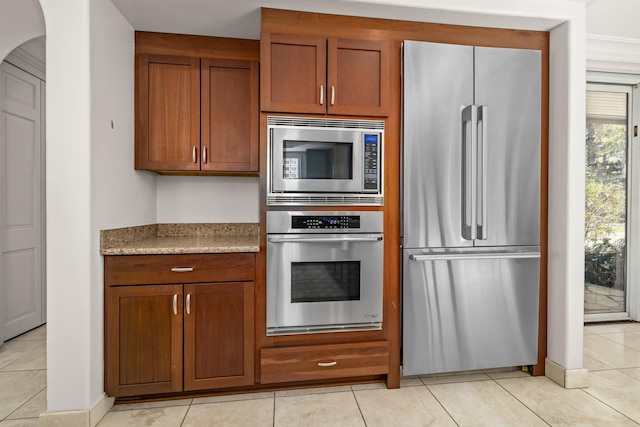 kitchen featuring light tile patterned floors, stainless steel appliances, light stone counters, and brown cabinetry