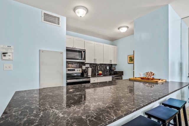 kitchen with stainless steel appliances, a kitchen breakfast bar, dark stone counters, white cabinets, and backsplash