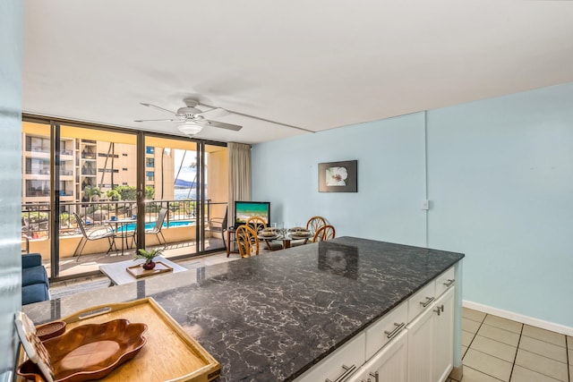 kitchen featuring a wall of windows, ceiling fan, dark stone countertops, light tile flooring, and white cabinetry