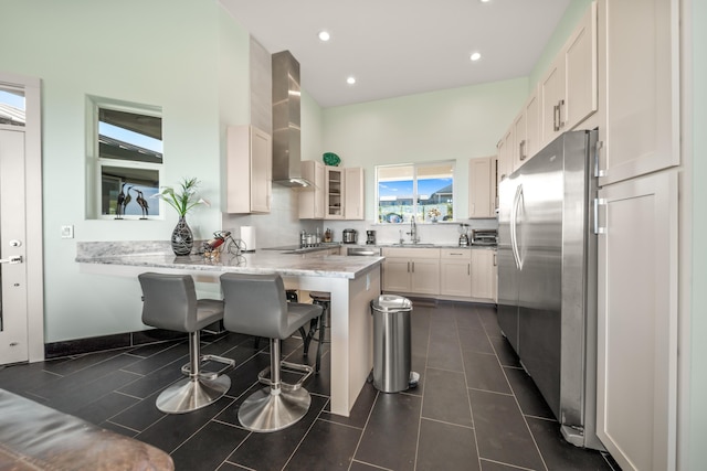 kitchen featuring a breakfast bar, wall chimney range hood, sink, white cabinetry, and stainless steel refrigerator