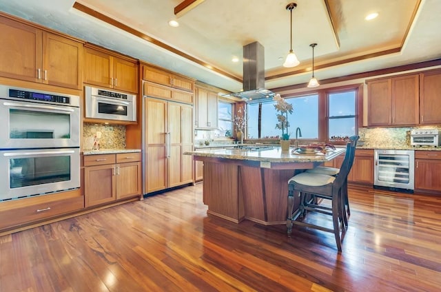 kitchen with island exhaust hood, a tray ceiling, wine cooler, light stone countertops, and double oven