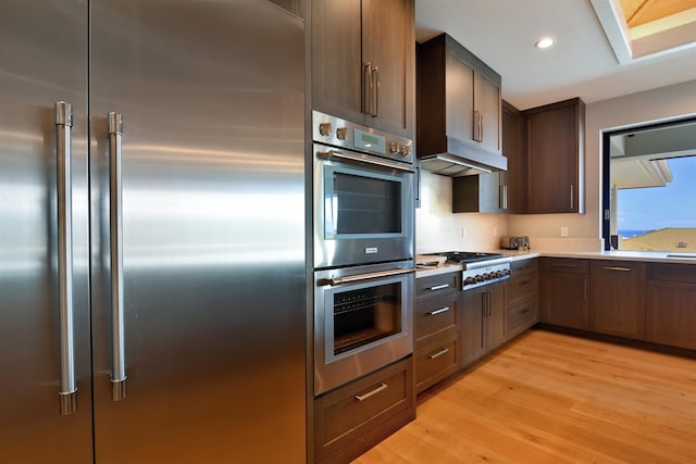 kitchen with a skylight, light wood-type flooring, dark brown cabinets, stainless steel appliances, and range hood