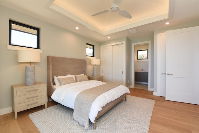 bedroom with ceiling fan, a tray ceiling, light hardwood / wood-style floors, and ensuite bathroom