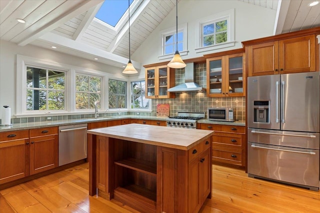 kitchen with stainless steel appliances, wall chimney exhaust hood, hanging light fixtures, a kitchen island, and light wood-type flooring