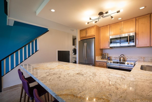 kitchen with light brown cabinets, a breakfast bar area, light stone counters, stacked washer / dryer, and stainless steel appliances