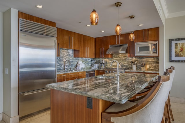 kitchen featuring built in appliances, extractor fan, a sink, dark stone counters, and pendant lighting