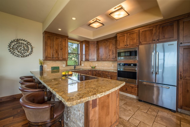 kitchen featuring sink, built in appliances, a kitchen breakfast bar, a raised ceiling, and kitchen peninsula